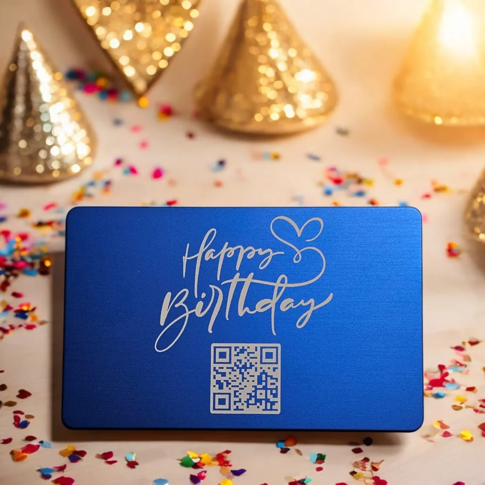 Blaue Aluminiumkarte mit "Happy Birthday"-Gravur und QR-Code, über welchen eine persönliche Botschaft aufgerufen werden kann Die Metallkarte ist umgeben von goldenen Partyhüten und buntem Konfetti.