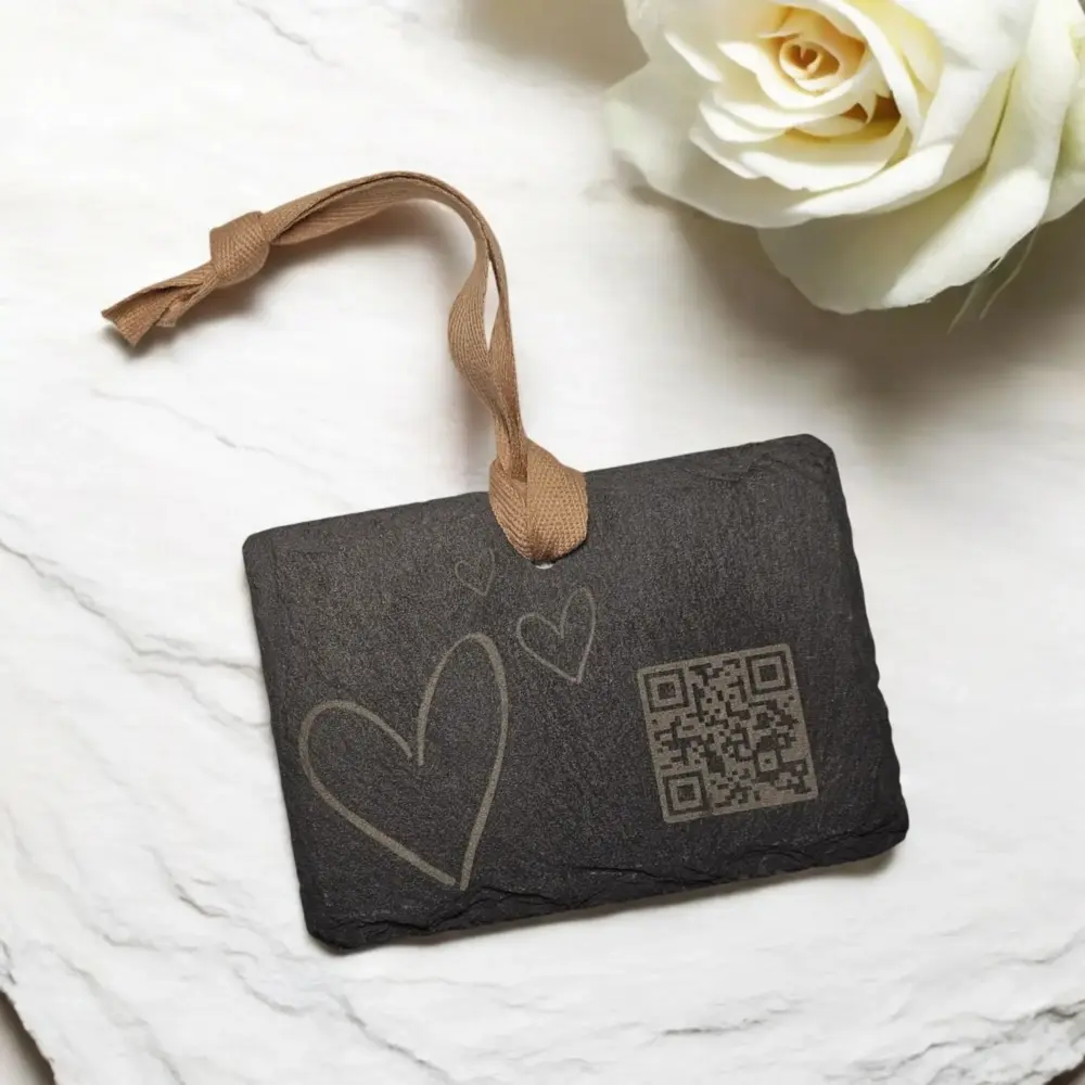 Dunkler Schieferanhänger mit Herzgravur und QR-Code auf hellem Untergrund – liebevoll inszeniert mit weißen Blumen
