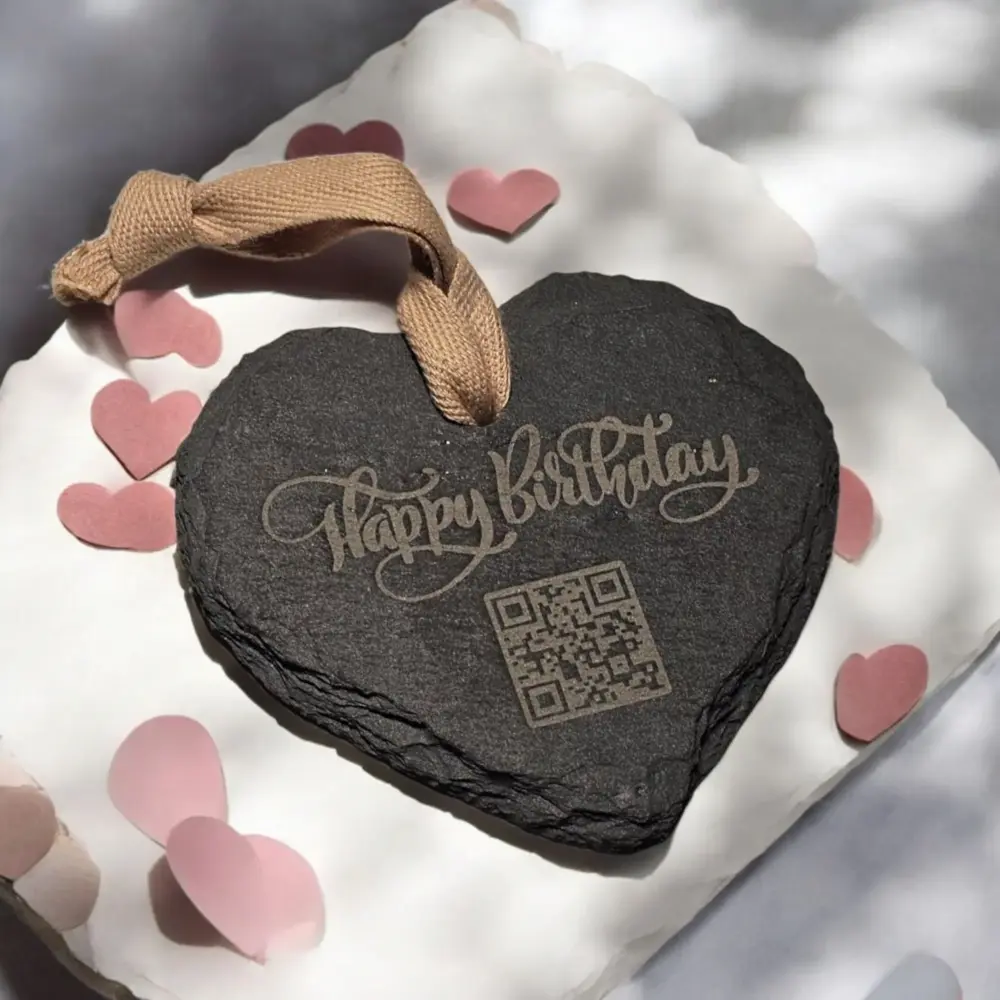 Herzförmiger Schieferanhänger mit QR-Code und Happy-Birthday-Gravur, umgeben von rosa Papierherzen