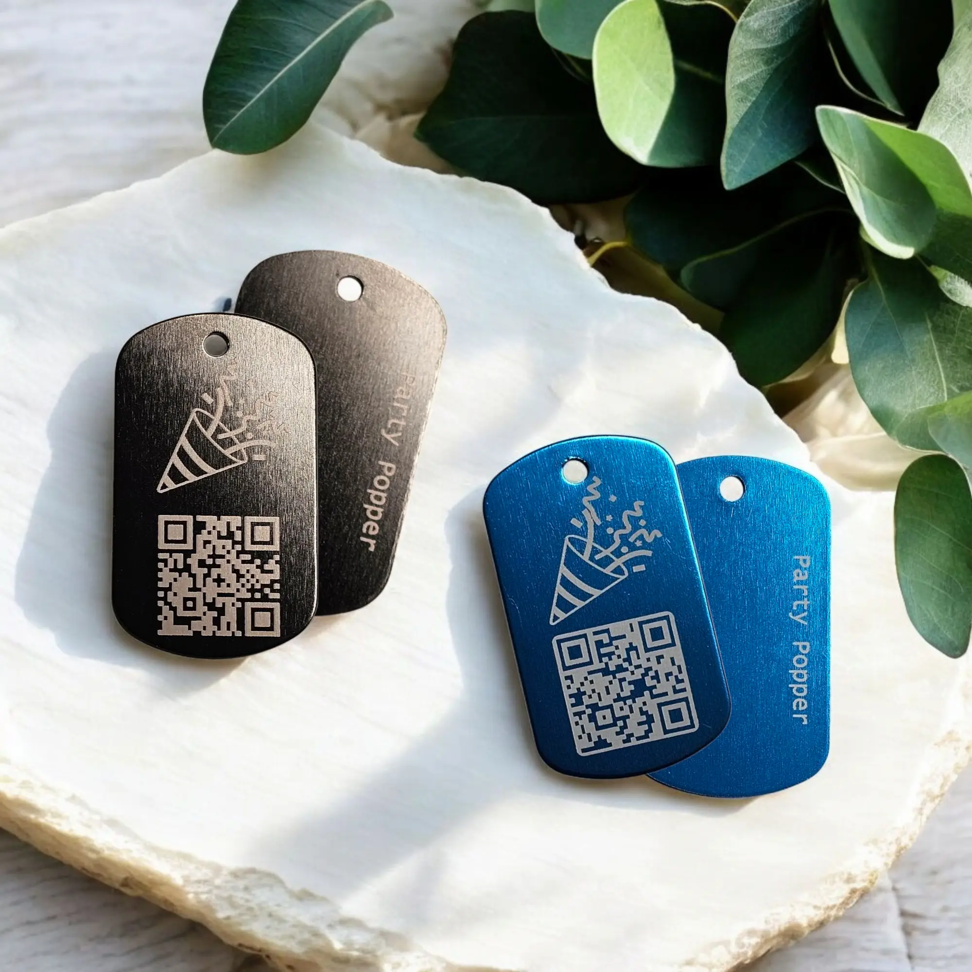 Vier Party Popper Anhänger mit QR-Code und graviertem Schriftzug auf schwarzem und blauem Aluminium – modernes Geschenk mit digitaler Botschaft.